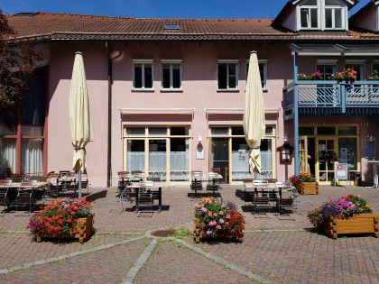 Vorderansicht vom Café, Stühle und Tische vor einem zweistöckigem Haus mit lachsfarbenem verputzten Wänden und einem hellblauen Balkon.