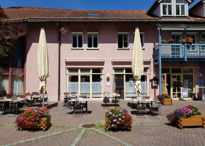Vorderansicht vom Café, Stühle und Tische vor einem zweistöckigem Haus mit lachsfarbenem verputzten Wänden und einem hellblauen Balkon.
